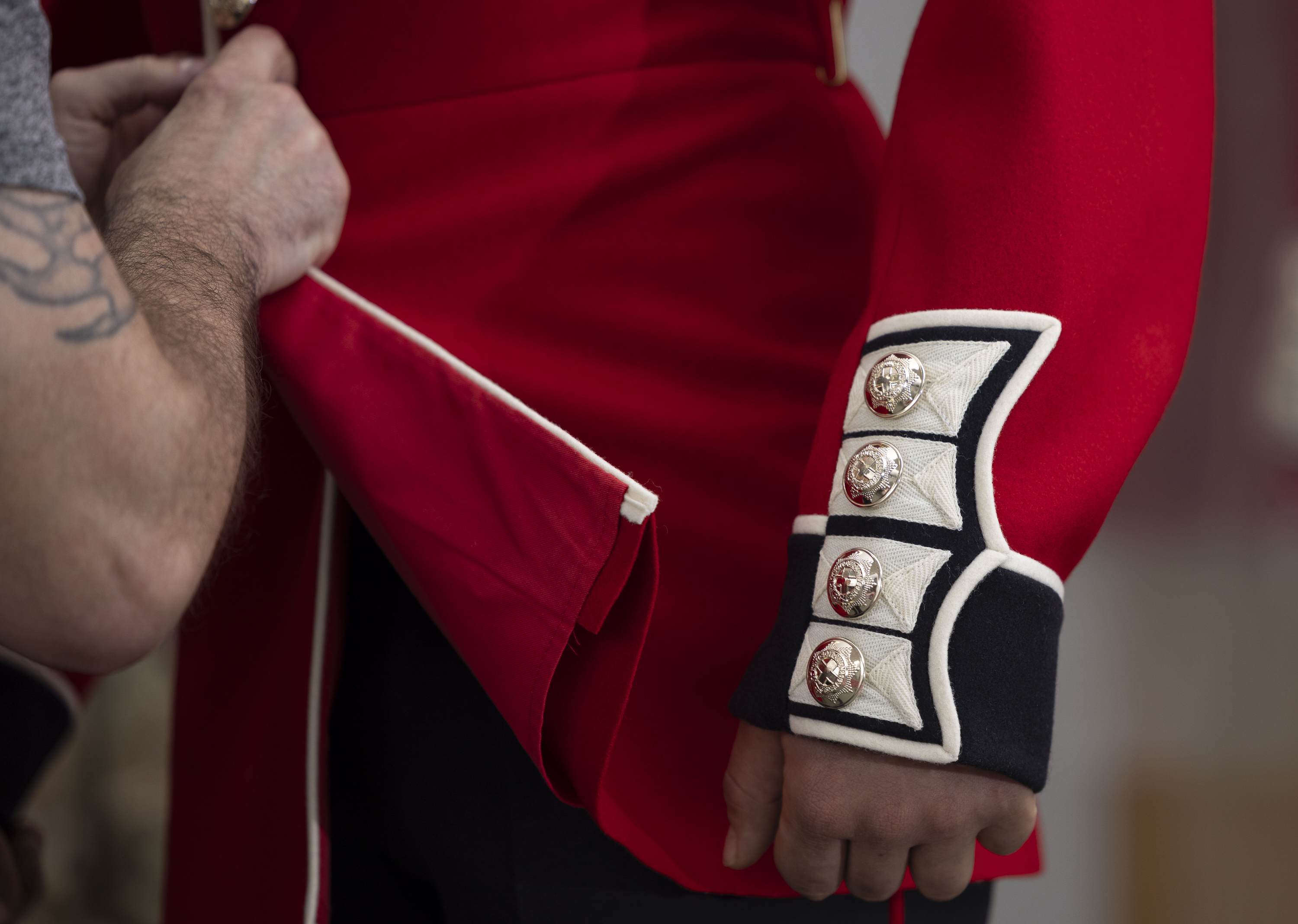 This image shows someone buttoning a red tunic as the ceremonial dress of the Household Division. The jacket features coldstream guards cap badge imprinted on shiny, metallic buttons going down the cuff of the tunic.