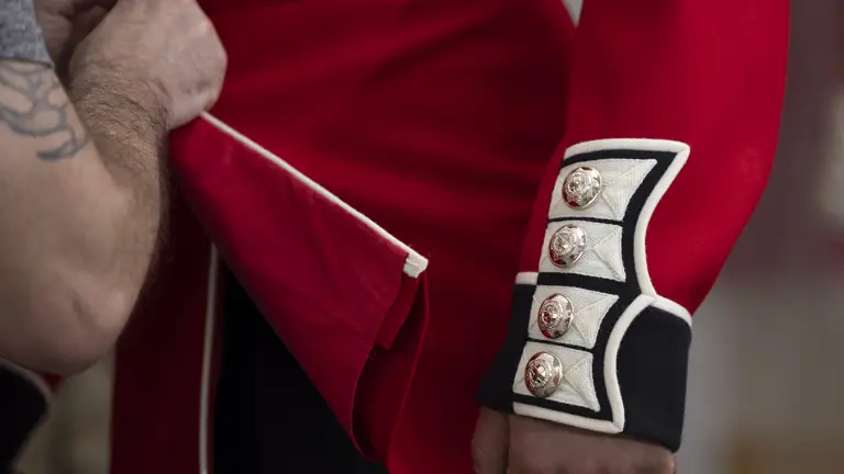 This image shows someone buttoning a red tunic as the ceremonial dress of the Household Division. The jacket features coldstream guards cap badge imprinted on shiny, metallic buttons going down the cuff of the tunic.