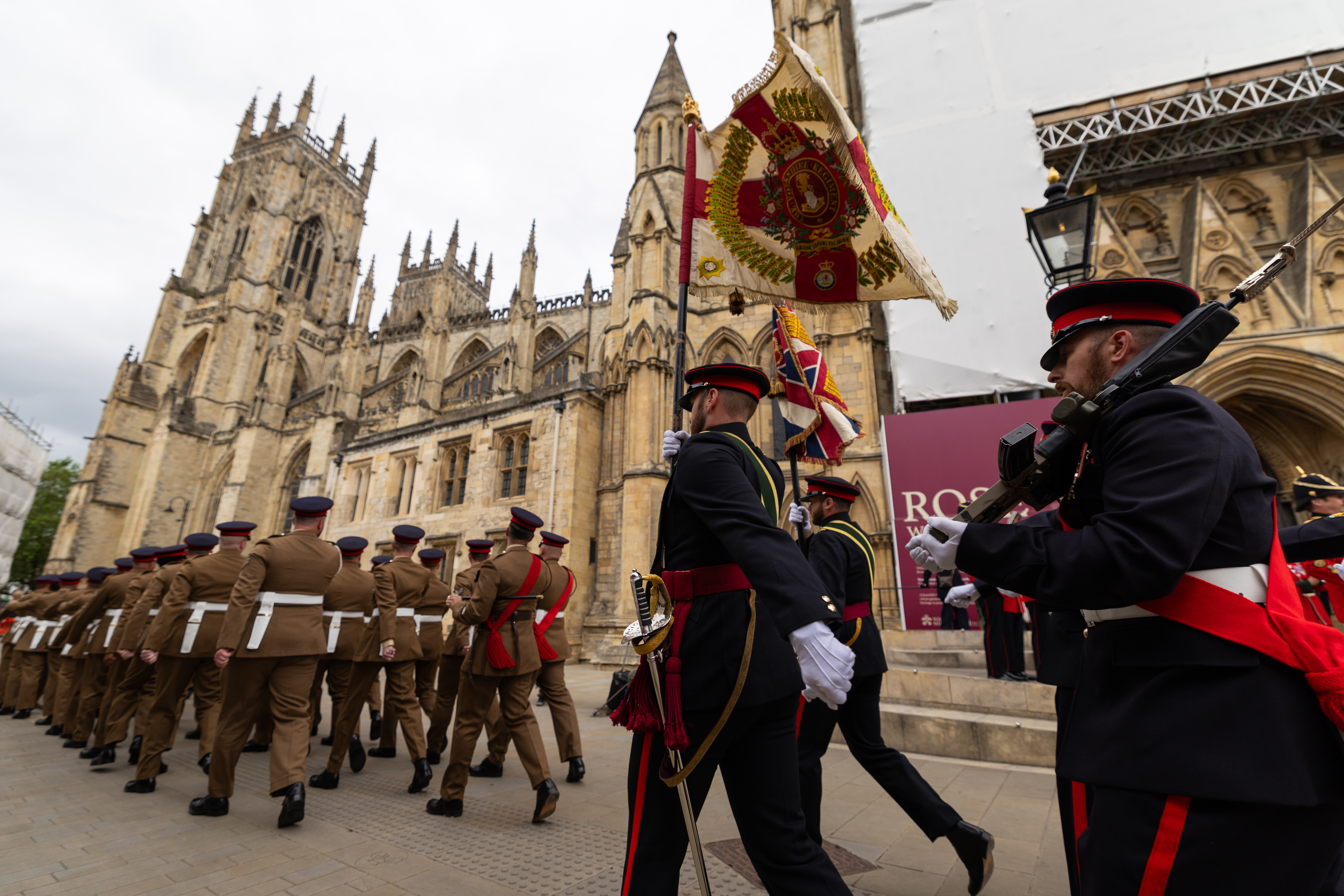 A ceremonial parade takes place outside the historic York Minster. Individuals in formal military attire march in formation, some carrying colorful flags and instruments. The grand gothic architecture of the cathedral serves as a backdrop.