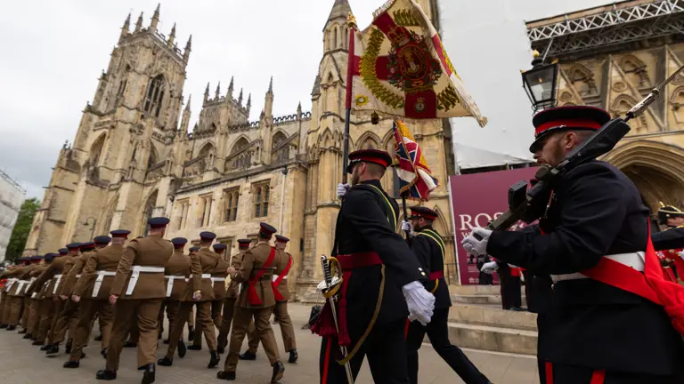 A ceremonial parade takes place outside the historic York Minster. Individuals in formal military attire march in formation, some carrying colorful flags and instruments. The grand gothic architecture of the cathedral serves as a backdrop.