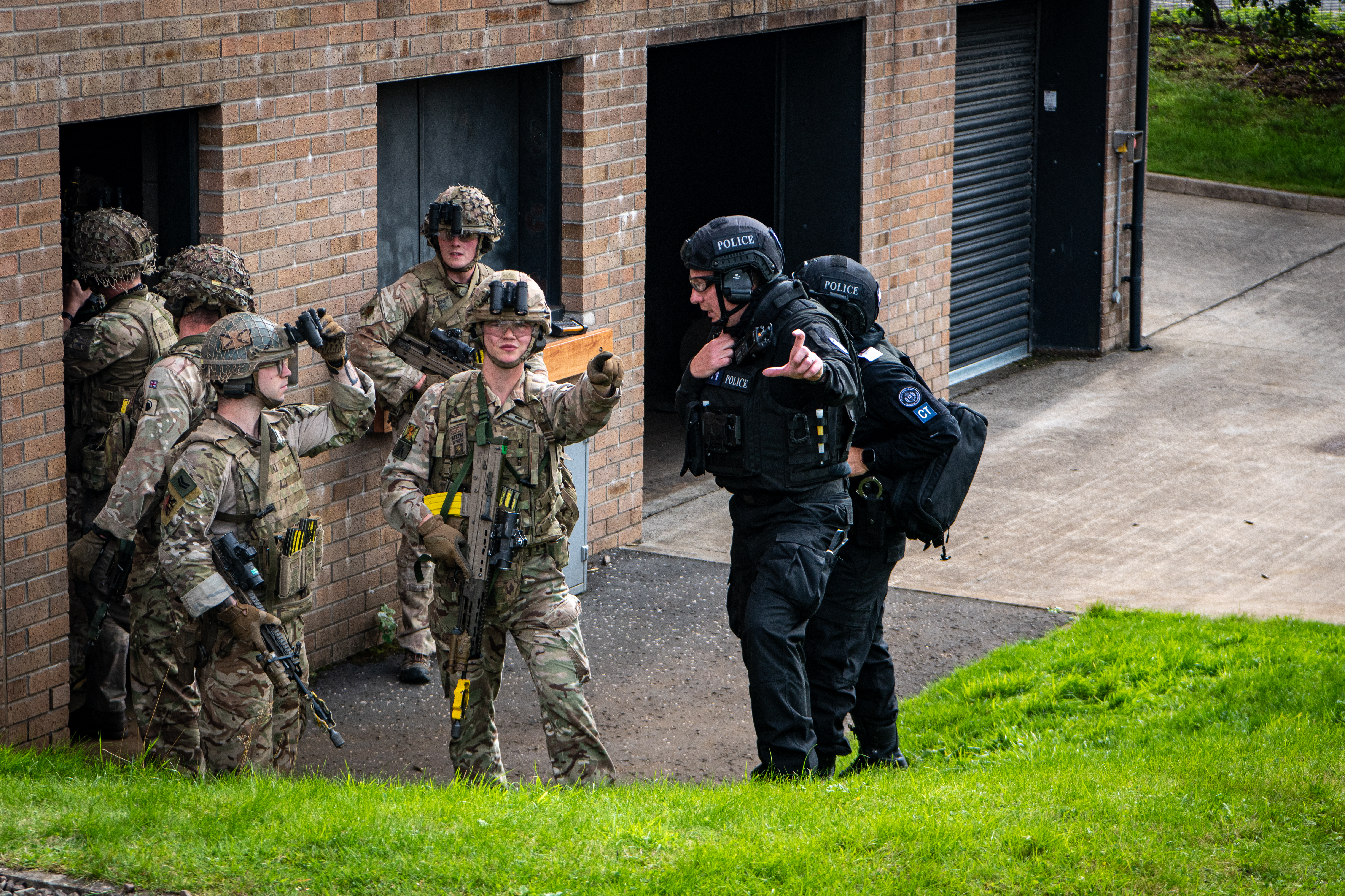 A group of people in military camouflage and police uniforms collaborate outside a brick building. Some are pointing and appear to be engaged in a tactical exercise.