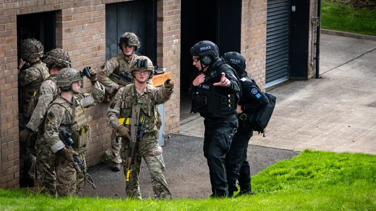 A group of people in military camouflage and police uniforms collaborate outside a brick building. Some are pointing and appear to be engaged in a tactical exercise.