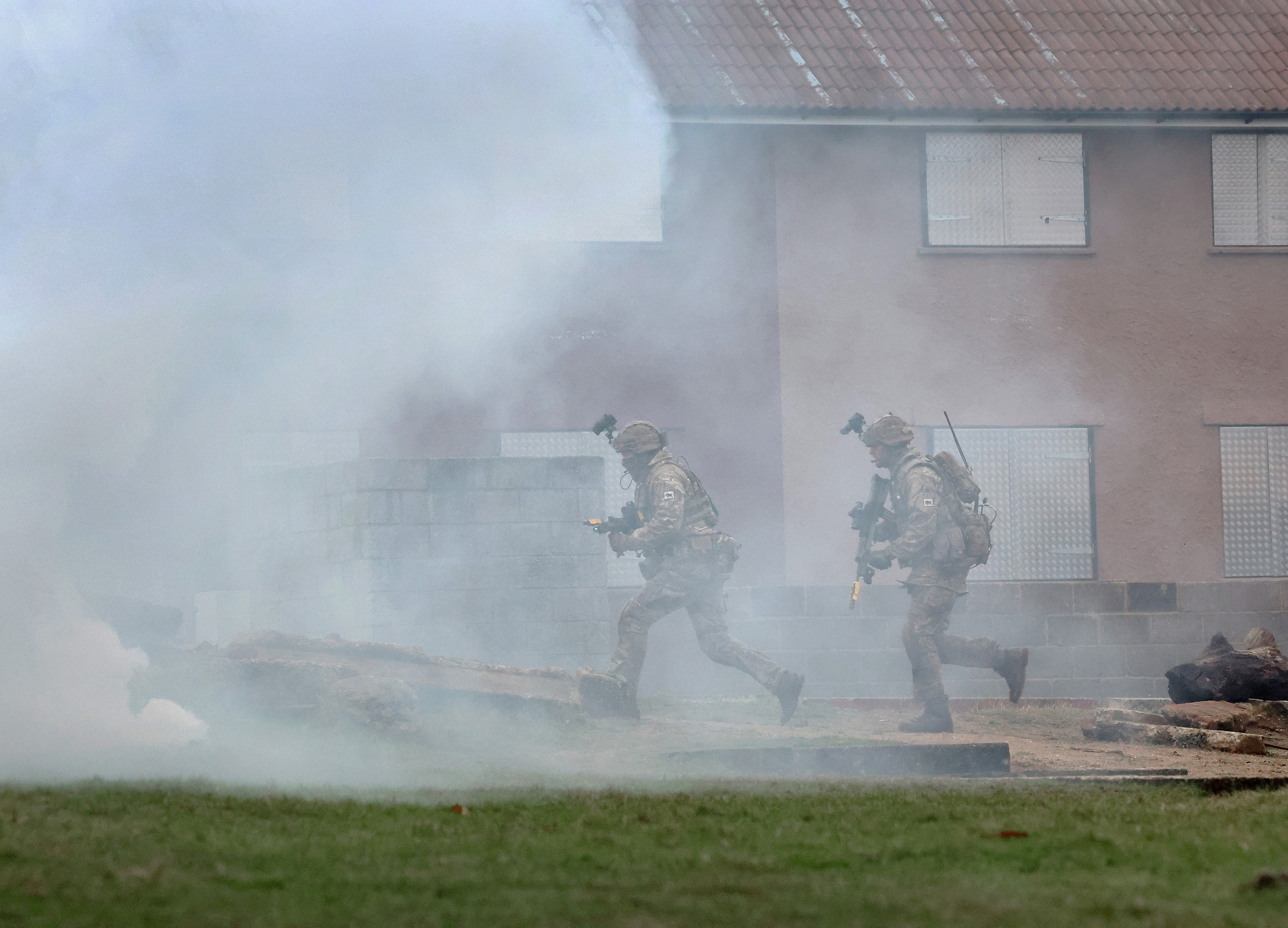 Soldiers wearing camouflage uniforms and carrying rifles run through some white smoke in front of houses.