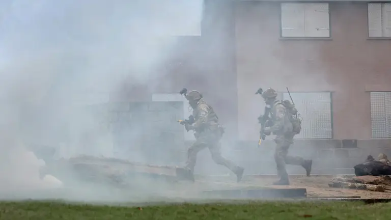 Soldiers wearing camouflage uniforms and carrying rifles run through some white smoke in front of houses.