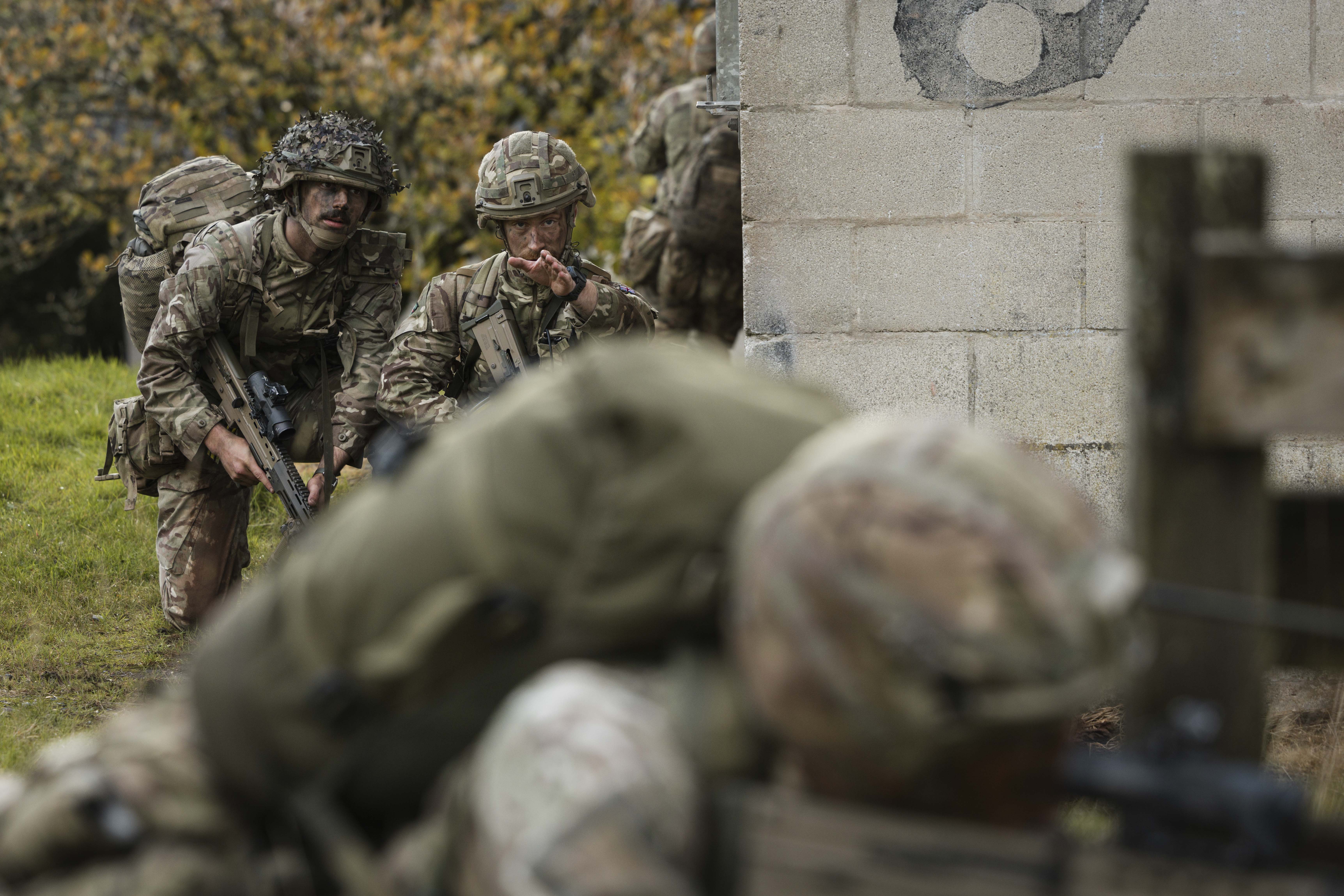 Soldiers in camouflage uniforms and helmets navigate a grassy area beside a stone wall. One soldier gestures, indicating a tactical maneuver. 