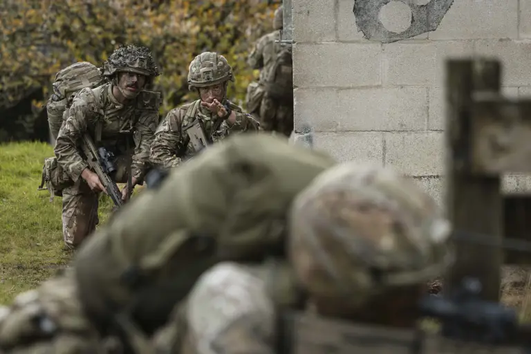 Soldiers in camouflage uniforms and helmets navigate a grassy area beside a stone wall. One soldier gestures, indicating a tactical maneuver.