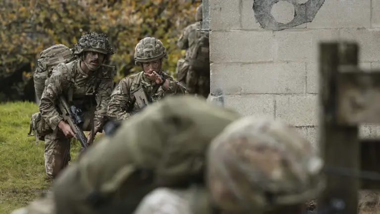 Soldiers in camouflage uniforms and helmets navigate a grassy area beside a stone wall. One soldier gestures, indicating a tactical maneuver.