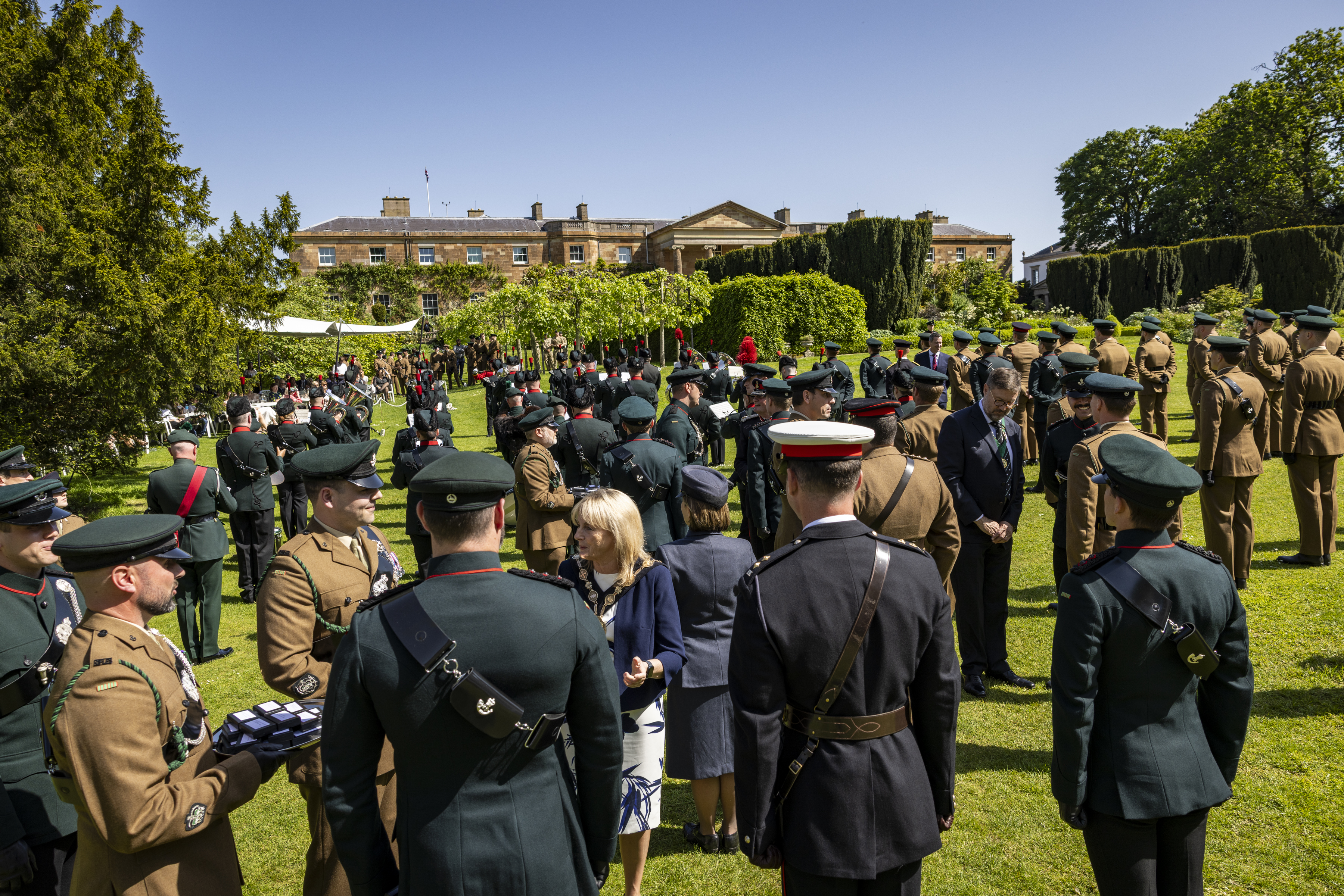 A large group of people in military uniforms gather in a garden outside a large historic building. The scene includes various individuals engaged in conversation and a presentation or ceremony is taking place. Lush greenery and a clear sky provide a scenic backdrop.