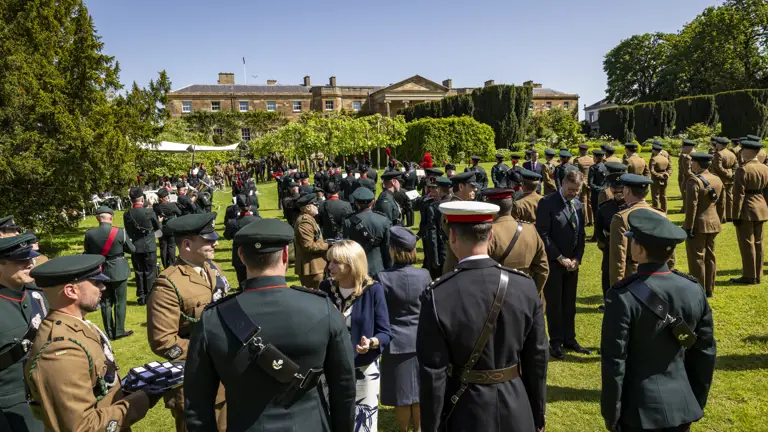 A large group of people in military uniforms gather in a garden outside a large historic building. The scene includes various individuals engaged in conversation and a presentation or ceremony is taking place. Lush greenery and a clear sky provide a scenic backdrop.