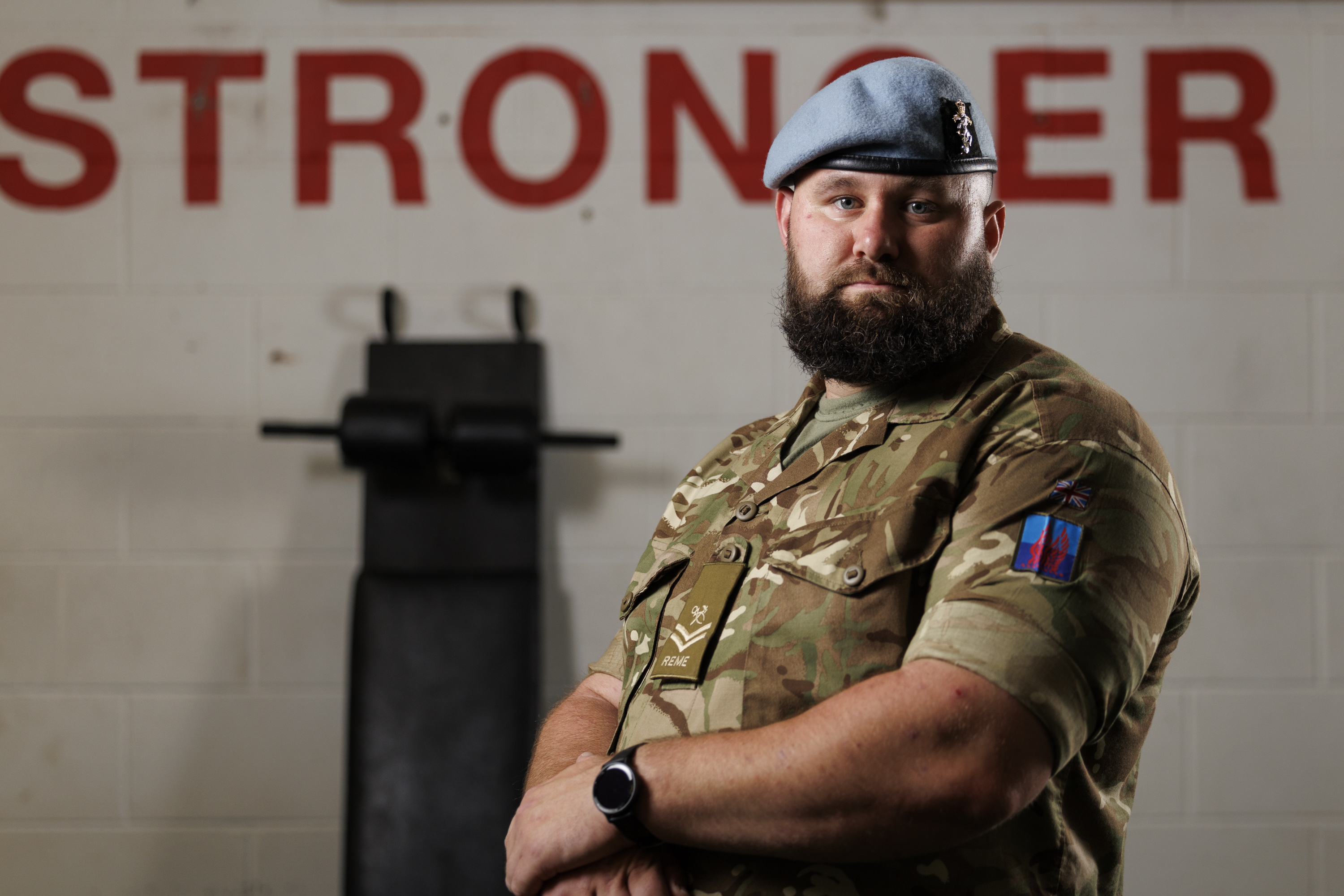 A bearded man in a military uniform and blue beret stands confidently in front of a wall with 