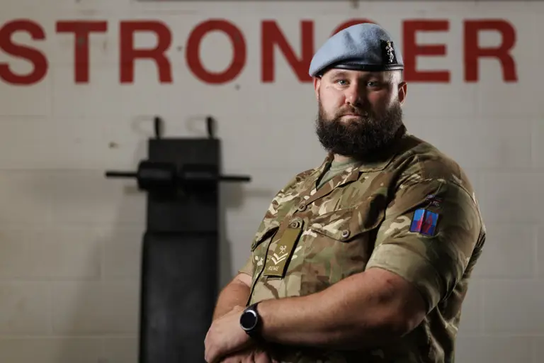 A bearded man in a military uniform and blue beret stands confidently in front of a wall with
