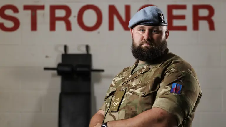 A bearded man in a military uniform and blue beret stands confidently in front of a wall with