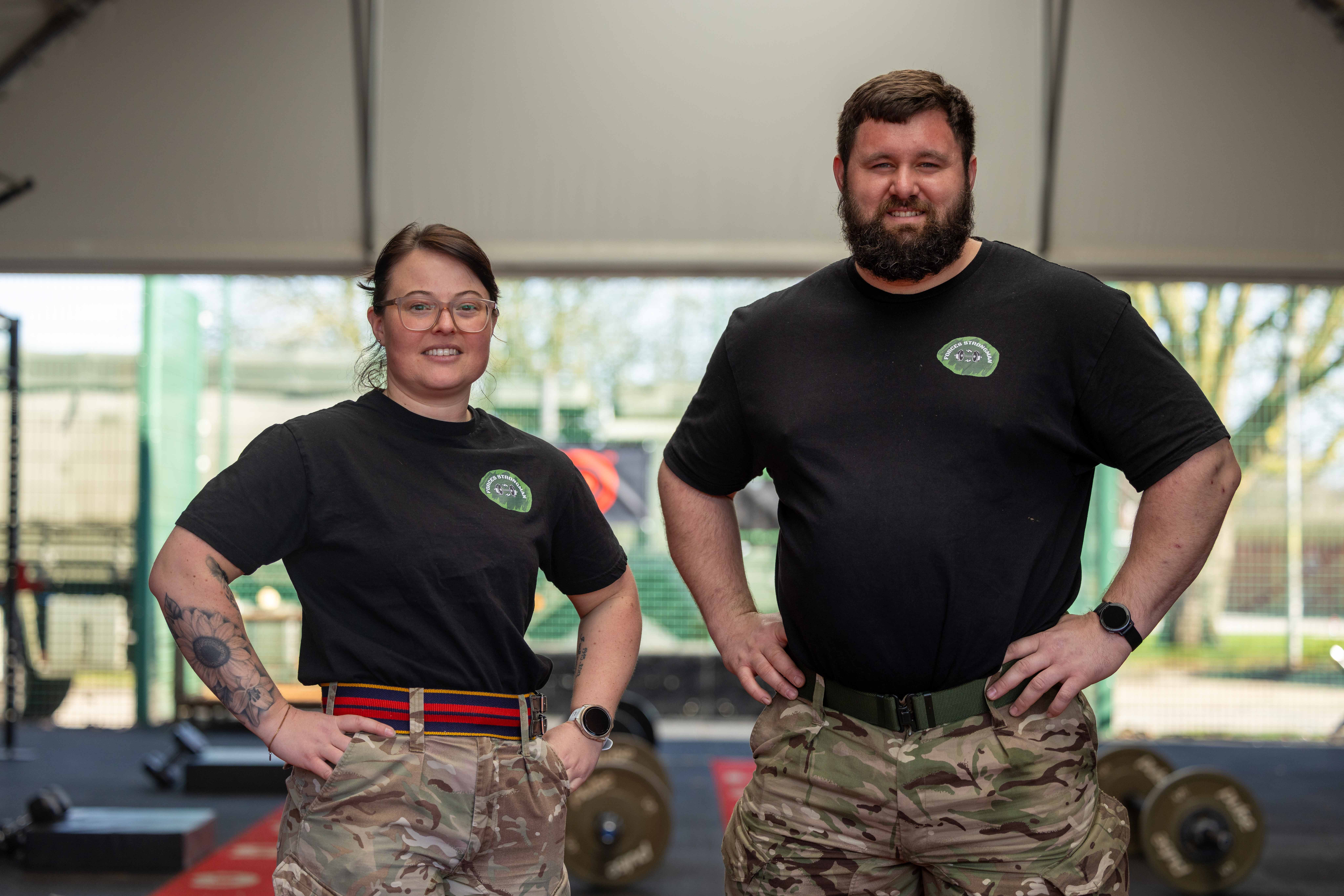 Two individuals in black shirts and camouflage pants stand confidently in a gym. Weights are visible in the background, suggesting a focus on fitness.