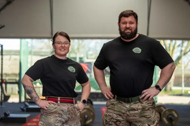 Two individuals in black shirts and camouflage pants stand confidently in a gym. Weights are visible in the background, suggesting a focus on fitness.