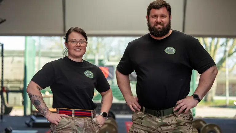 Two individuals in black shirts and camouflage pants stand confidently in a gym. Weights are visible in the background, suggesting a focus on fitness.