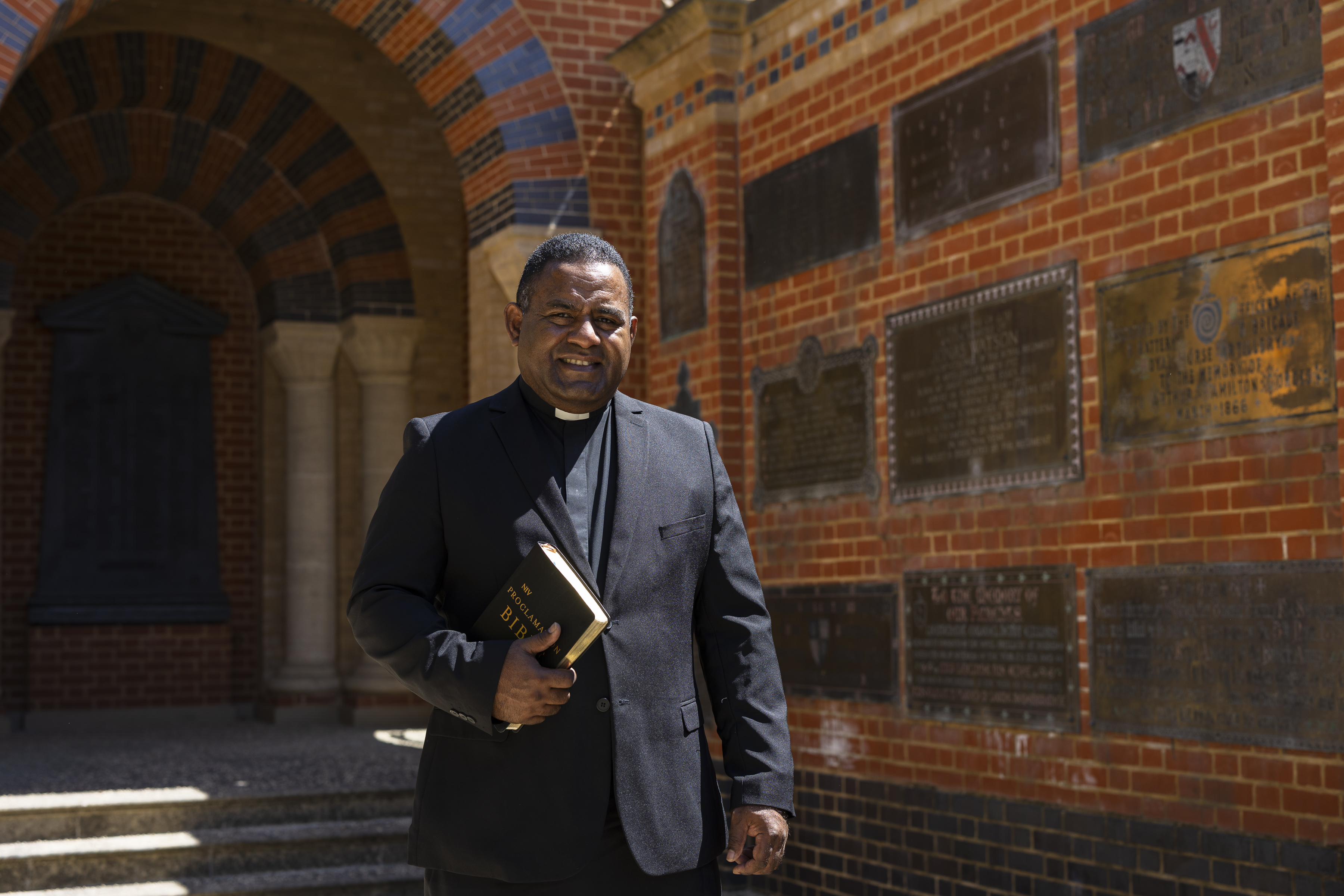 Clergyman in black suit holding a Bible standing in front of a brick wall with commemorative plaques.