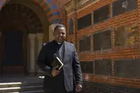 Clergyman in black suit holding a Bible standing in front of a brick wall with commemorative plaques.