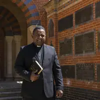 Clergyman in black suit holding a Bible standing in front of a brick wall with commemorative plaques.