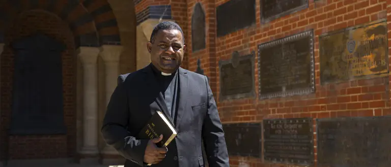 Clergyman in black suit holding a Bible standing in front of a brick wall with commemorative plaques.