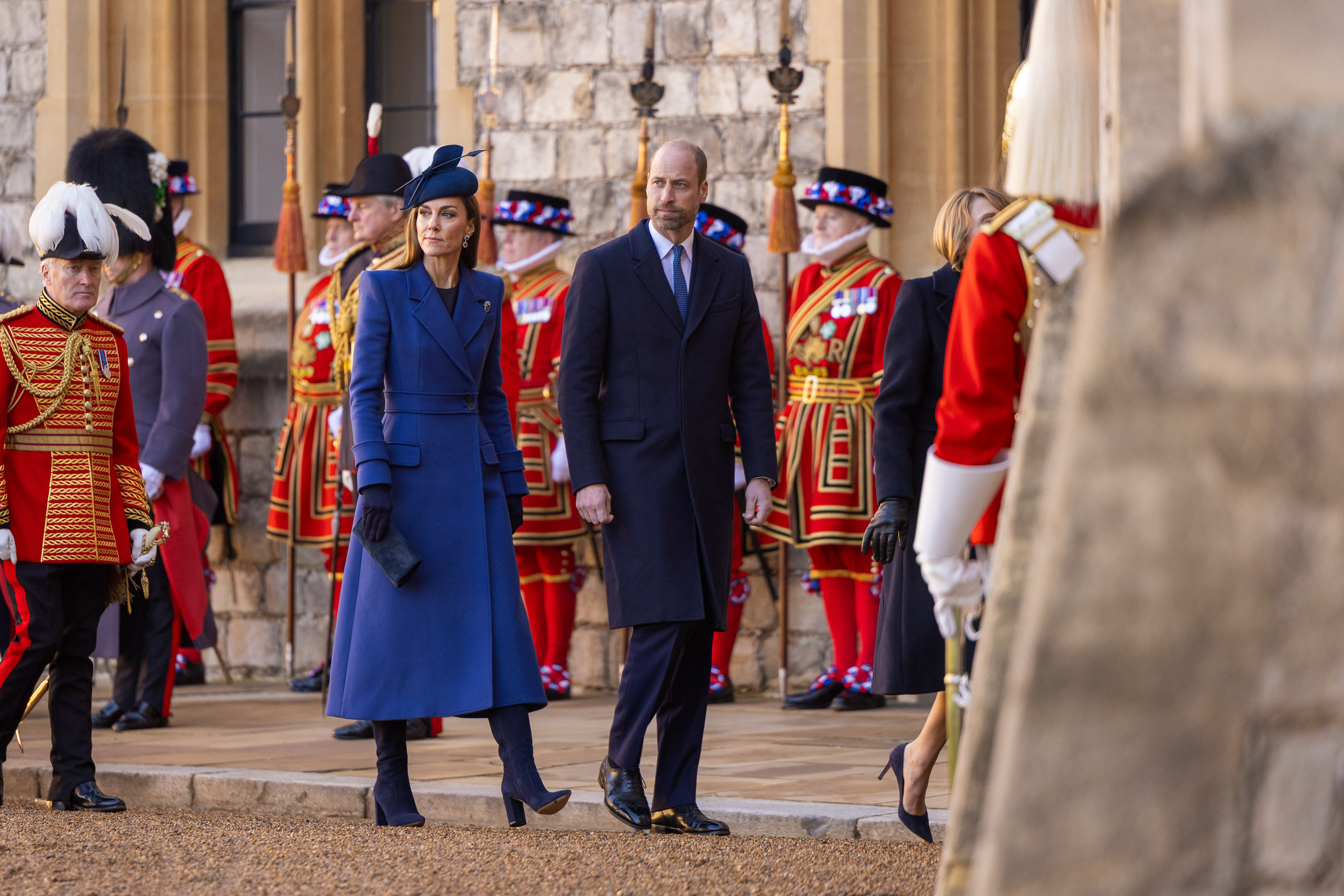 A man and woman in elegant coats walk together outside a historic building. Beefeaters in red uniforms stand in the background, adding a formal touch.