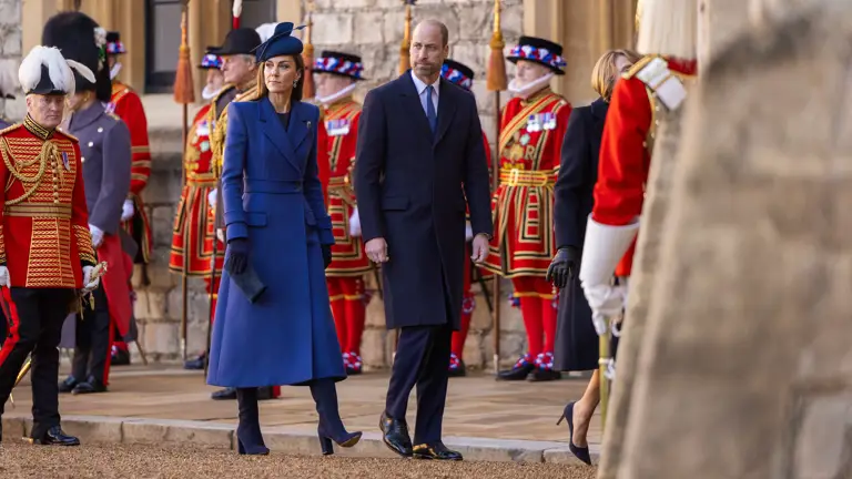 A man and woman in elegant coats walk together outside a historic building. Beefeaters in red uniforms stand in the background, adding a formal touch.