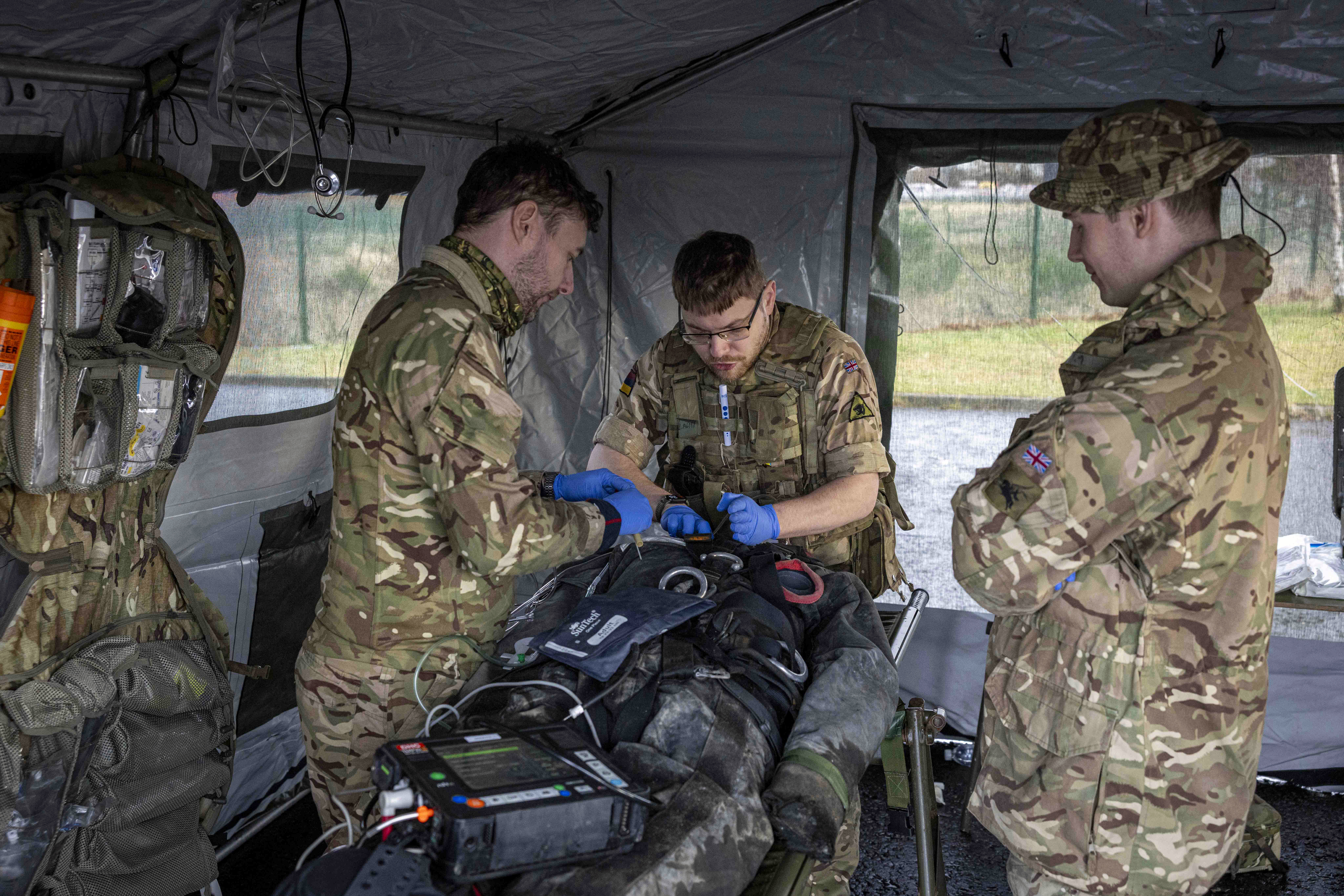 Three soldiers in camouflage uniforms provide medical treatment to a patient lying on a stretcher inside a field tent.