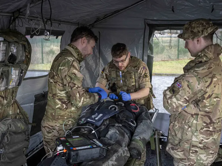 Three soldiers in camouflage uniforms provide medical treatment to a patient lying on a stretcher inside a field tent.