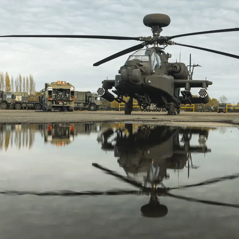 An Apache attack helicopter is parked on a tarmac beside a large puddle, reflecting the aircraft.