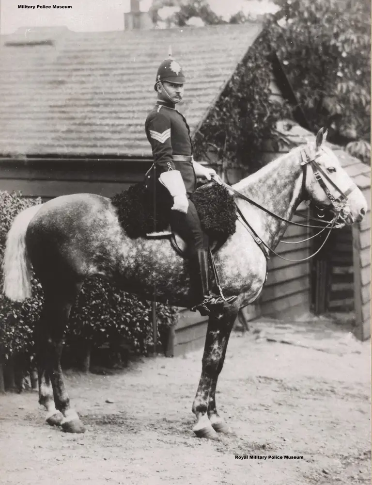 A mounted police officer in formal uniform sits confidently on a dappled horse. The background shows rustic wooden buildings and foliage, evoking a historical atmosphere.