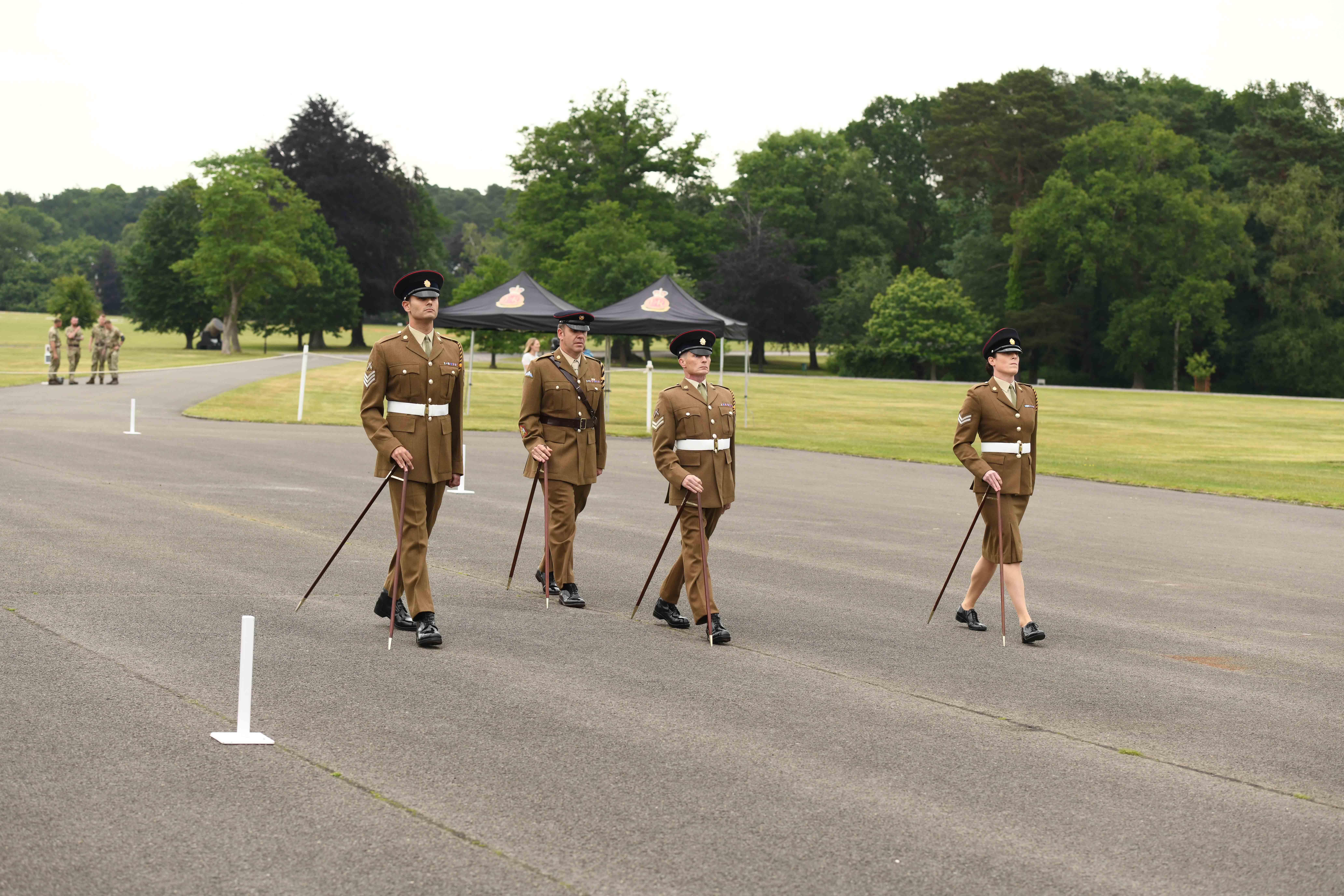 The make and one female British Army soldiers, wearing khaki dress uniform, are marching across a parade square using pace sticks. 