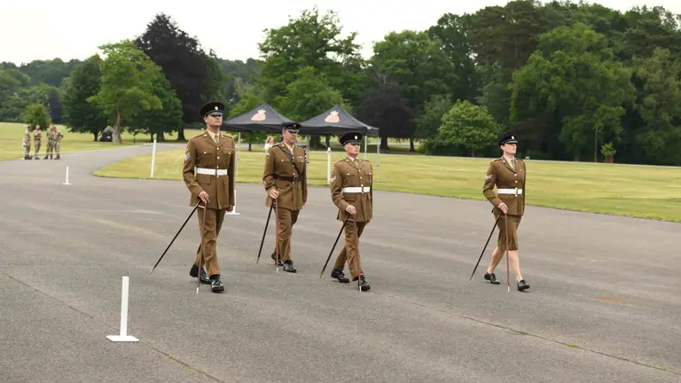 The make and one female British Army soldiers, wearing khaki dress uniform, are marching across a parade square using pace sticks.