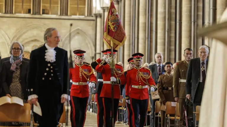 Men in red ceremonial uniforms carry a red and gold ceremonial flag through a cathedral.