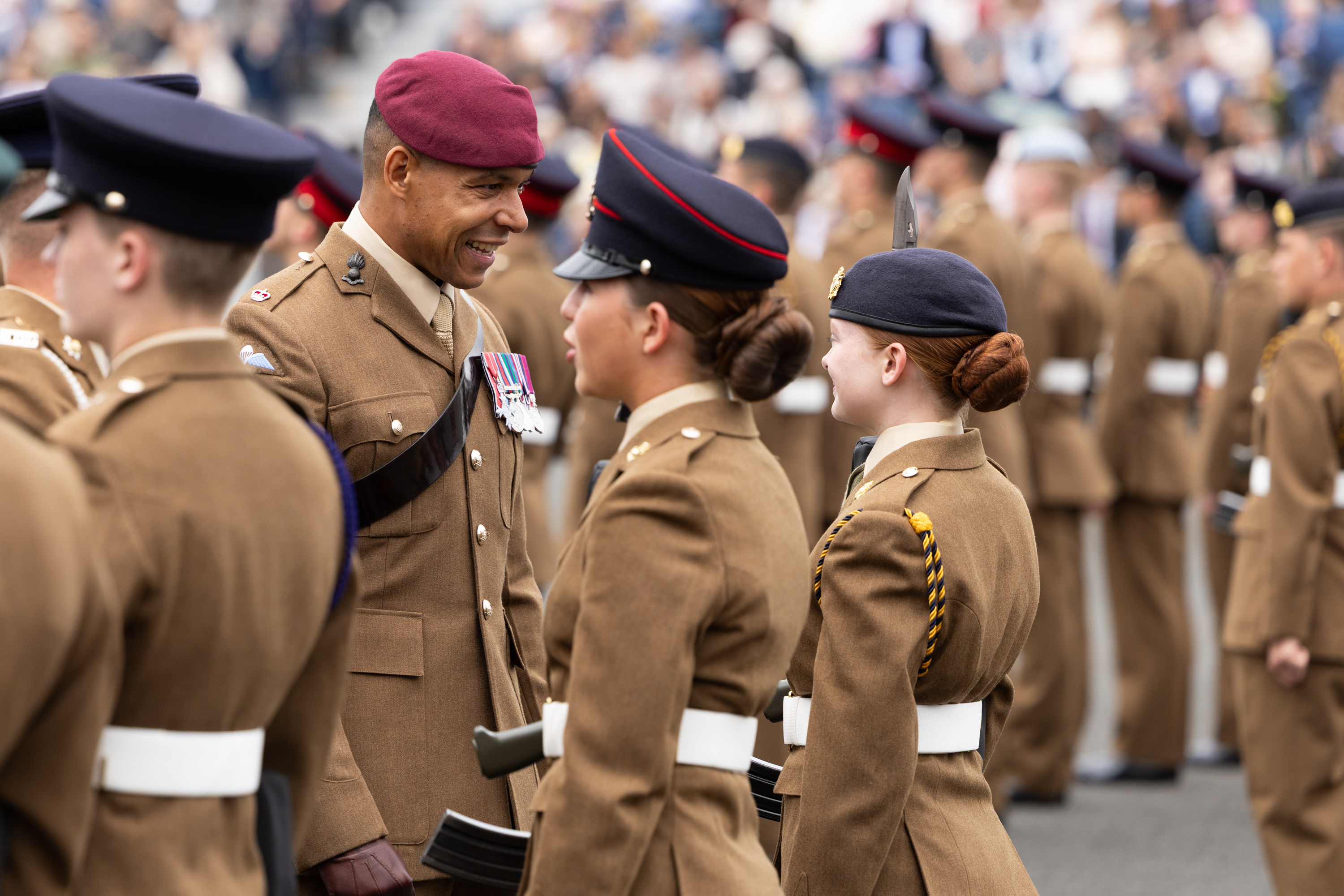 A group of uniformed soldiers stand in formation during a parade. One soldier wearing a red beret and decorated with medals interacts with others. The setting appears formal and ceremonial, with a blurred audience in the background.