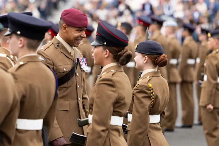 A group of uniformed soldiers stand in formation during a parade. One soldier wearing a red beret and decorated with medals interacts with others. The setting appears formal and ceremonial, with a blurred audience in the background.