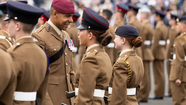 A group of uniformed soldiers stand in formation during a parade. One soldier wearing a red beret and decorated with medals interacts with others. The setting appears formal and ceremonial, with a blurred audience in the background.
