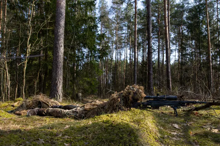 A camouflaged sniper lies on mossy ground taking aim with his rifle.