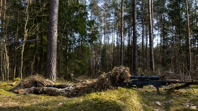 A camouflaged sniper lies on mossy ground taking aim with his rifle.