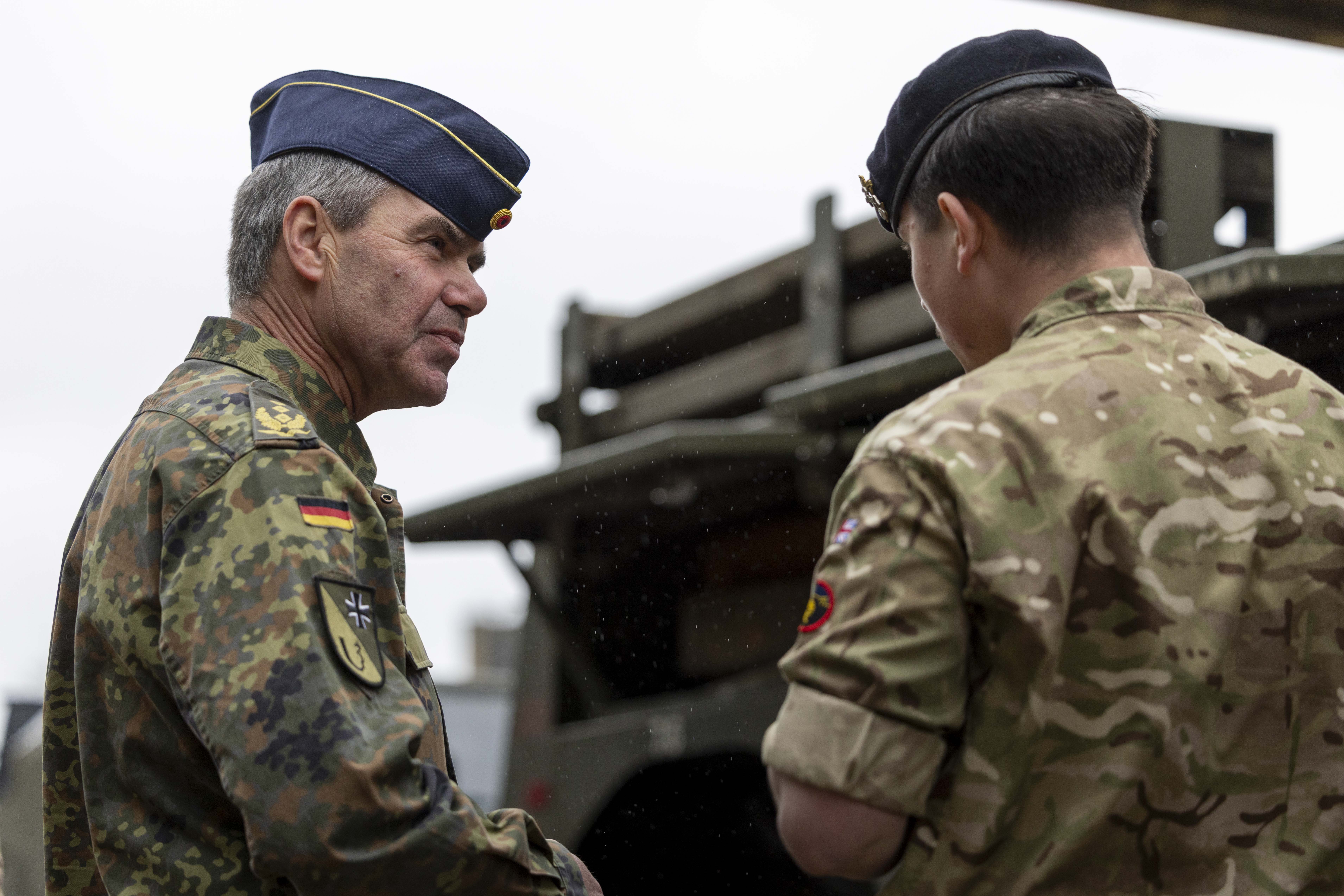Two military personnel in camouflage uniforms converse outdoors. One wears a peaked hat and the other a beret. They stand near a military vehicle.