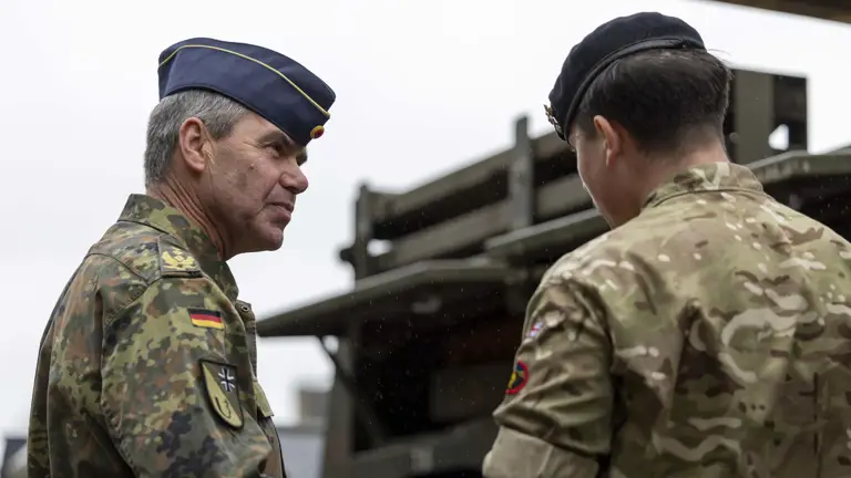 Two military personnel in camouflage uniforms converse outdoors. One wears a peaked hat and the other a beret. They stand near a military vehicle.