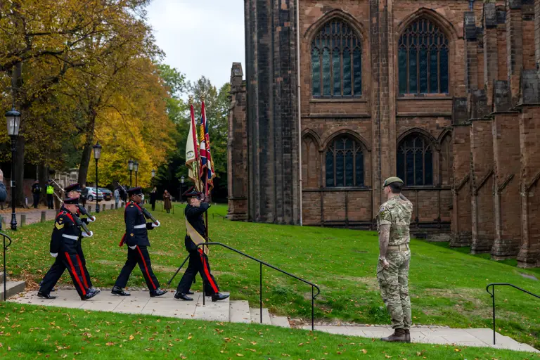Five soldiers wearing blues uniforms marching towards the cathedral with the colours flags being saluted by one man sat in the path wearing camouflage uniform.