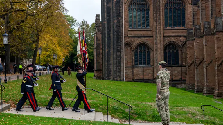 Five soldiers wearing blues uniforms marching towards the cathedral with the colours flags being saluted by one man sat in the path wearing camouflage uniform.