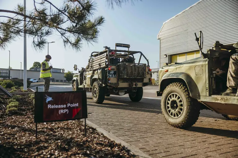 Military vehicles lined up at a designated release point outside a modern industrial building under clear skies.