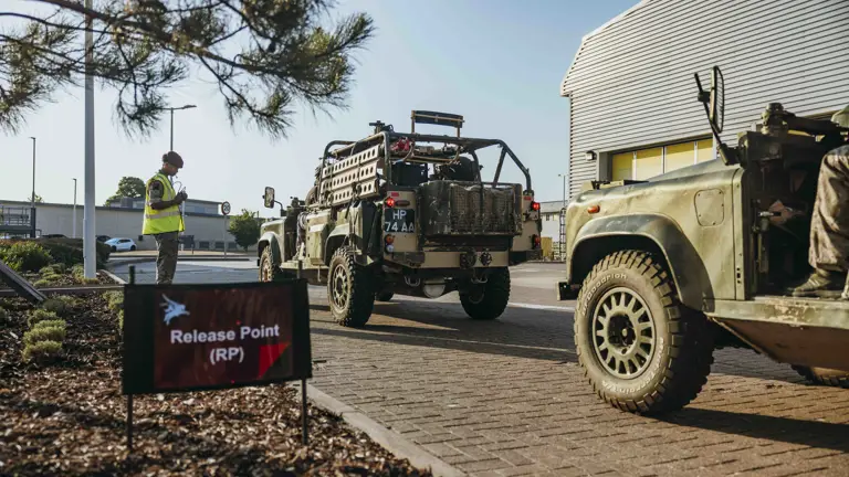 Military vehicles lined up at a designated release point outside a modern industrial building under clear skies.