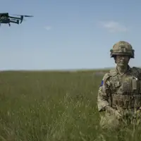 A soldier in camouflage gear stands in a grassy field, holding a controller. A green drone hovers in the sky nearby. The landscape is open with a clear blue sky.