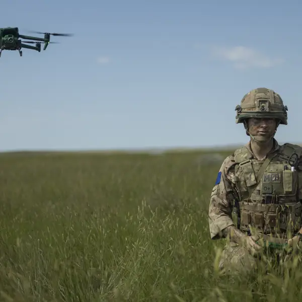 A soldier in camouflage gear stands in a grassy field, holding a controller. A green drone hovers in the sky nearby. The landscape is open with a clear blue sky.
