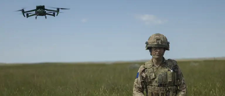 A soldier in camouflage gear stands in a grassy field, holding a controller. A green drone hovers in the sky nearby. The landscape is open with a clear blue sky.