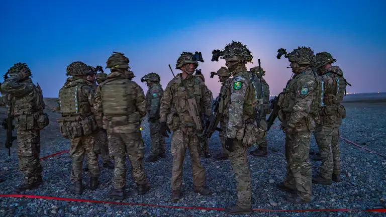 A group of 12 soldiers in camouflage uniform are seen in webbing and helmets stood waiting to start a training exercise in the darkness.