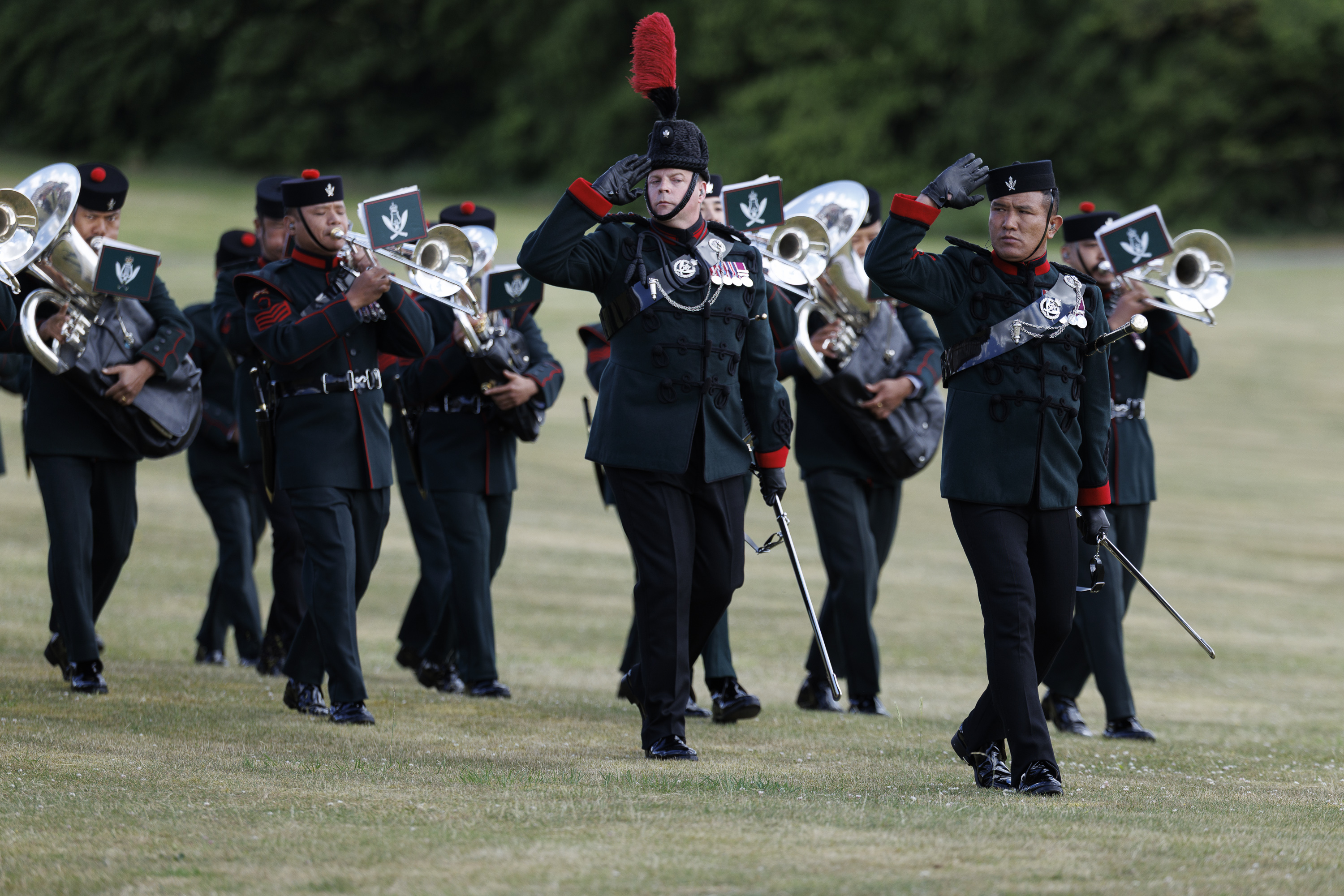 A marching band in dark green uniforms is performing on a grassy field. Several band members play brass instruments.