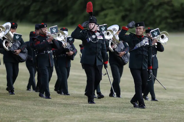 A marching band in dark green uniforms is performing on a grassy field. Several band members play brass instruments.