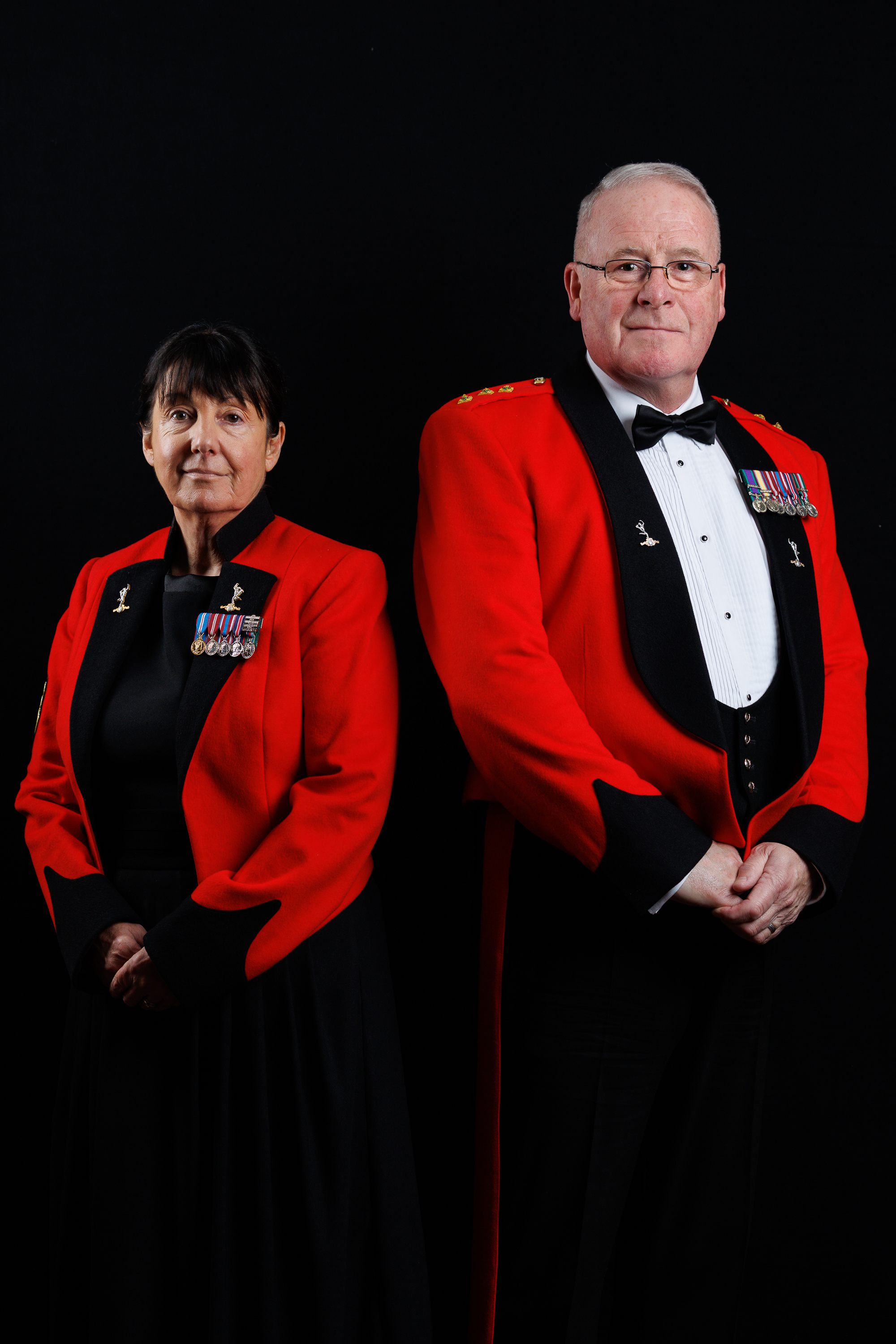 Two military personnel in formal red dress uniforms with medals stand against a black background.